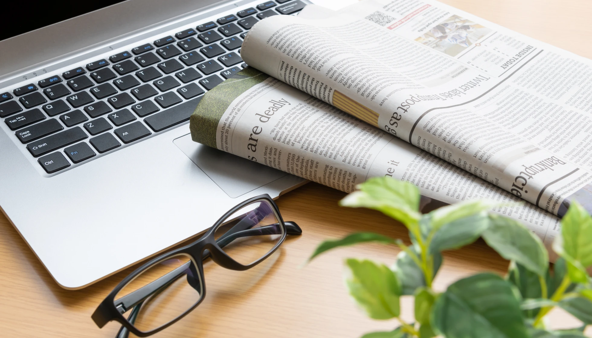デスクの上に置かれたノートパソコン、英字新聞、眼鏡と観葉植物