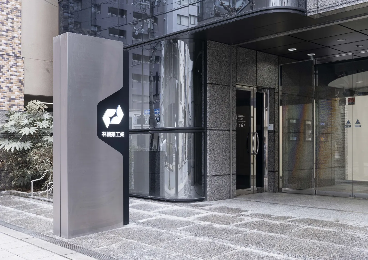 林純薬工業のロゴが入ったスタイリッシュな自立看板があるビルのエントランス。ガラス張りの外壁と石畳の地面