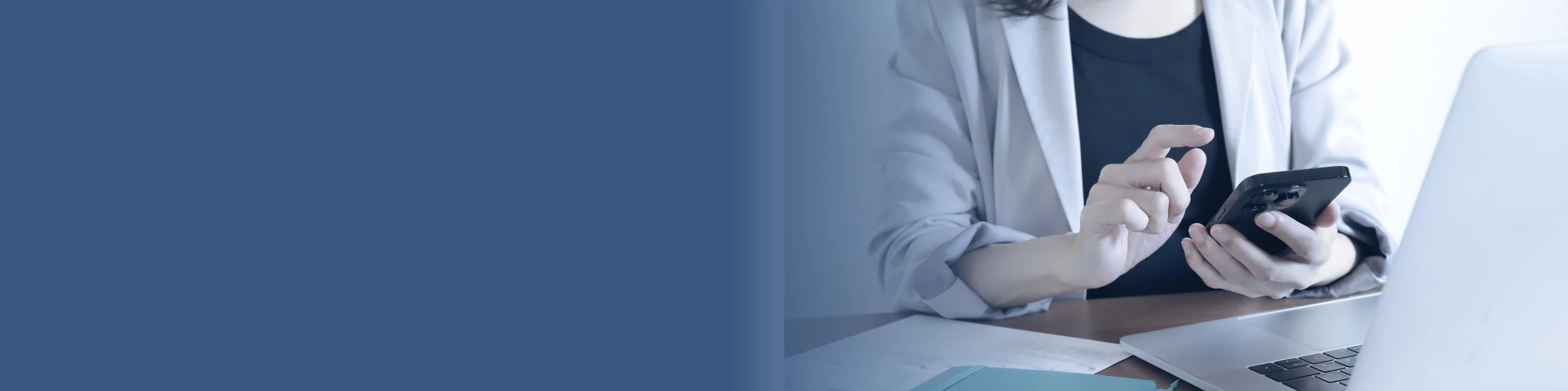 ノートパソコンの前でスマートフォンを操作する、オフィスカジュアルな服装の女性の手元