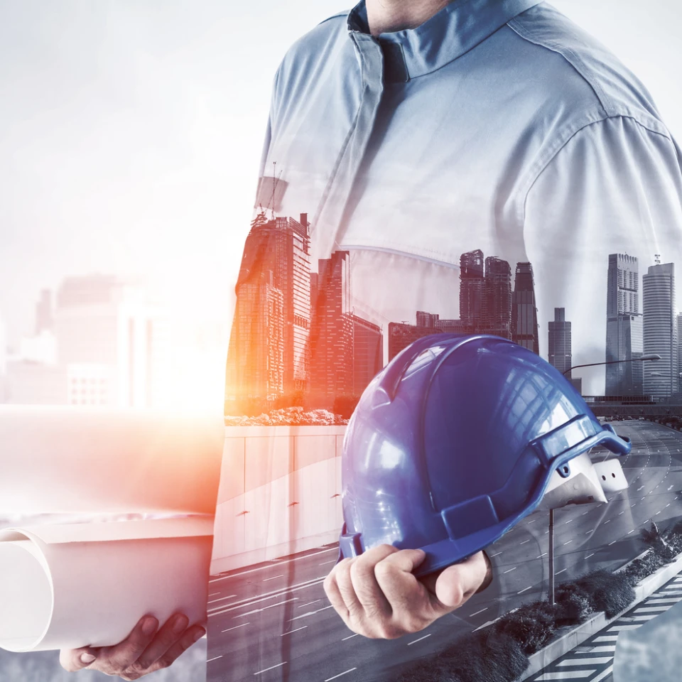 ヘルメットと図面を持つ作業員と、近代的なビル群や道路を重ねたダブル露出のイメージ