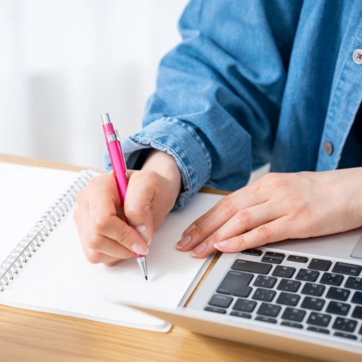 ノートパソコンの横で、ピンクのシャープペンシルを使いノートにメモを取るデスクワークの様子