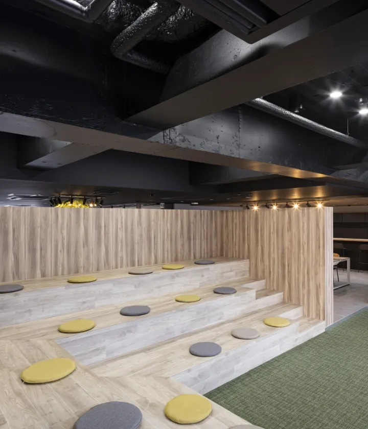 木目調の階段状の座席があるリラックススペース。黄色とグレーの円形クッションが並び、天井は黒でインダストリアルな雰囲気