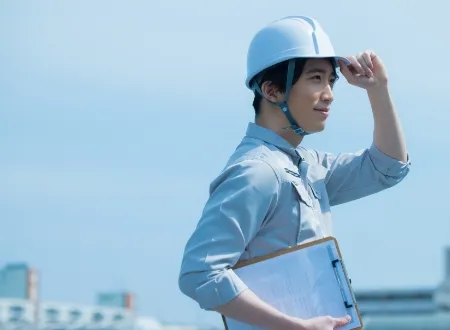 ヘルメットを被り、クリップボードを持って青空の下に立つ笑顔の男性作業員