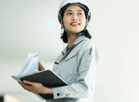 資料を確認しながら上方を向いて微笑む、ヘルメットと作業服姿の女性スタッフ