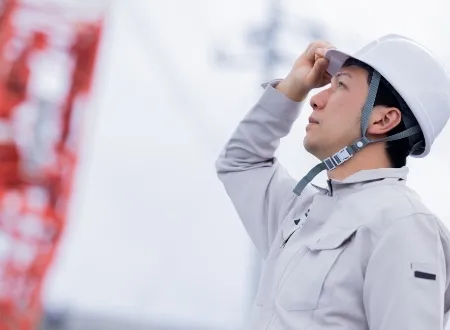 作業現場でヘルメットのつばに手をかけ、真剣な表情で空を見上げる男性作業員