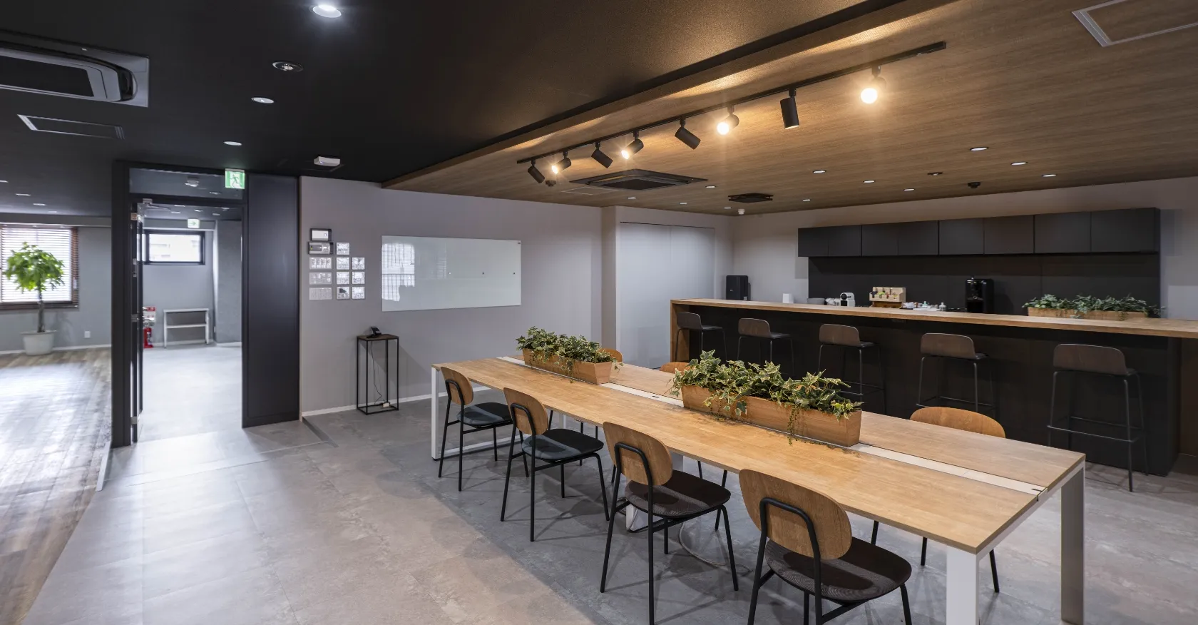 モダンなコワーキングスペース。長い木製テーブルに観葉植物、カウンター、そして黒い天井にスポットライト