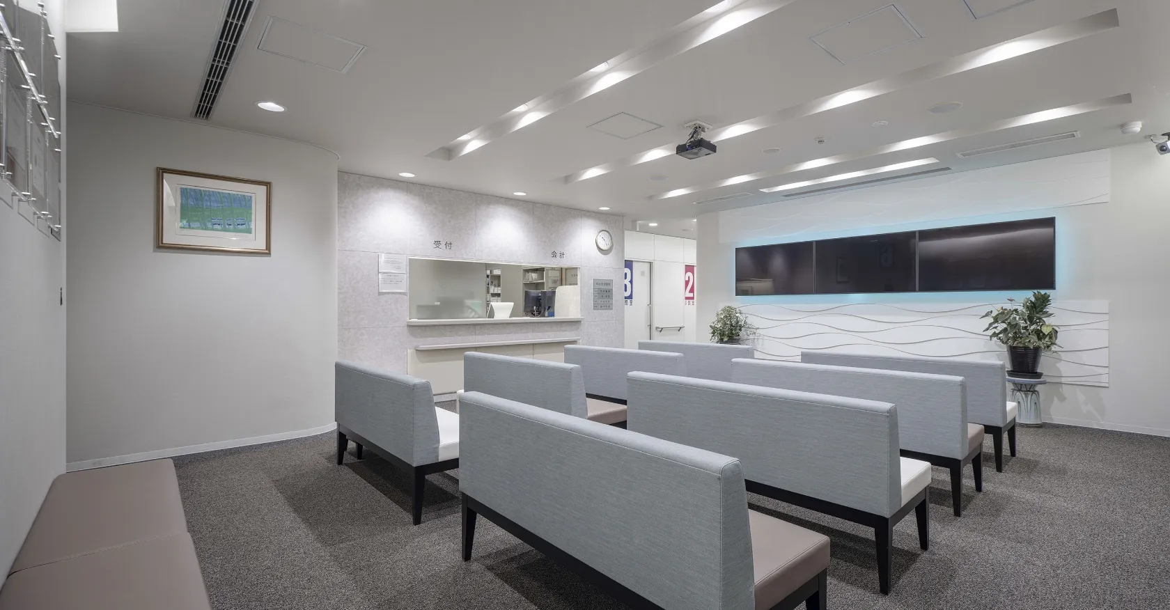 モダンで清潔感のあるクリニックの待合室。グレーのソファ、波模様の壁、大きなモニター、そして絵画が飾られている
