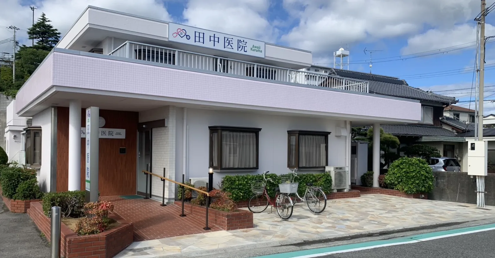 晴れた日の田中医院の外観。白い壁と淡いピンクのタイル、バルコニー、そして駐輪場には自転車が2台停まっている。