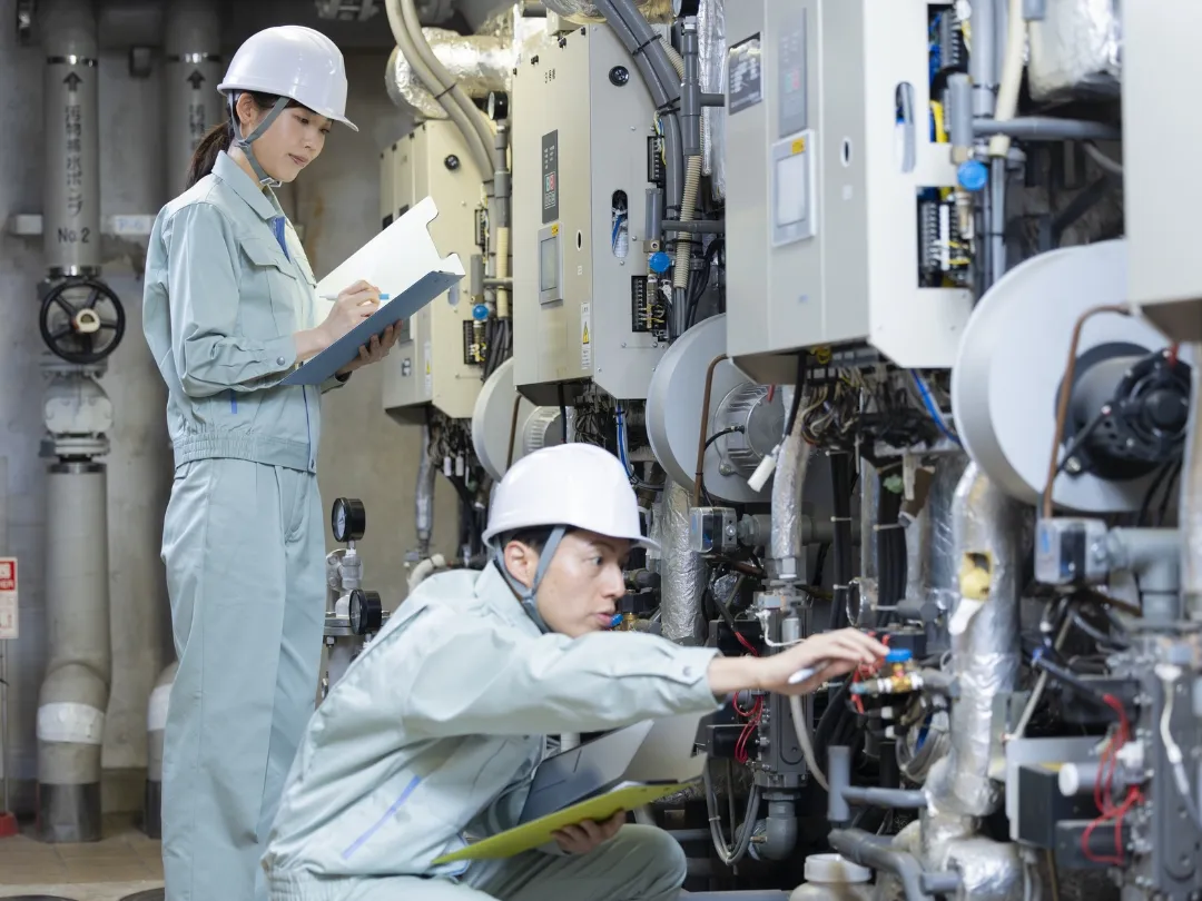 ヘルメットと作業着を着用した男女の技術者が、複雑な機械設備を点検している様子。一人はメモを取り、もう一人は機器を確認している