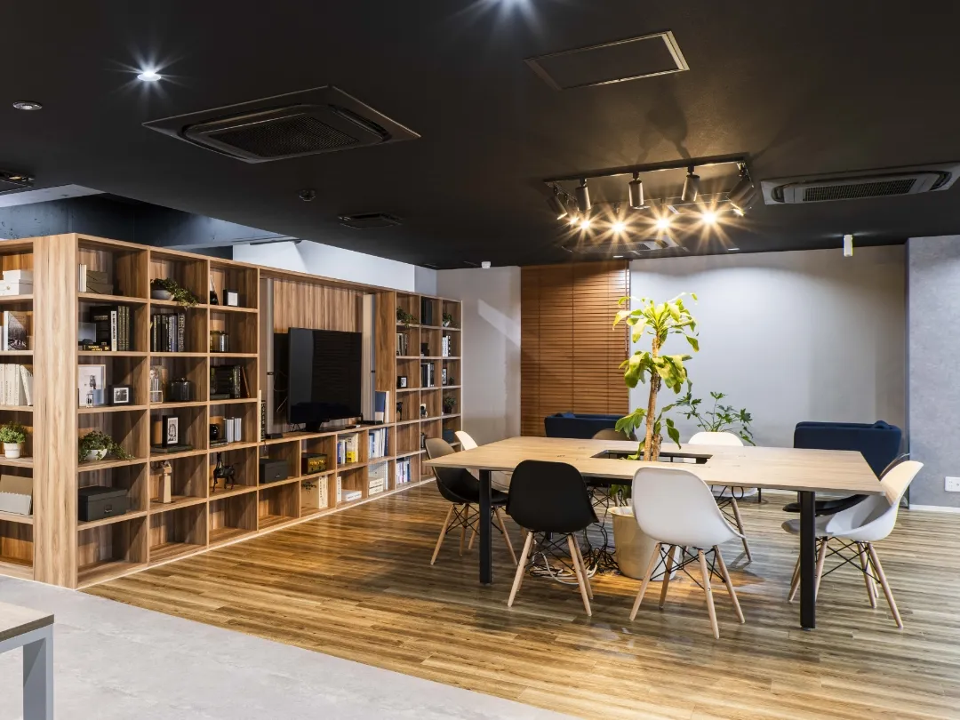 大きな木製の本棚があるオフィスのラウンジエリア。中央には植栽のあるテーブルとモダンな椅子があり、落ち着いた照明