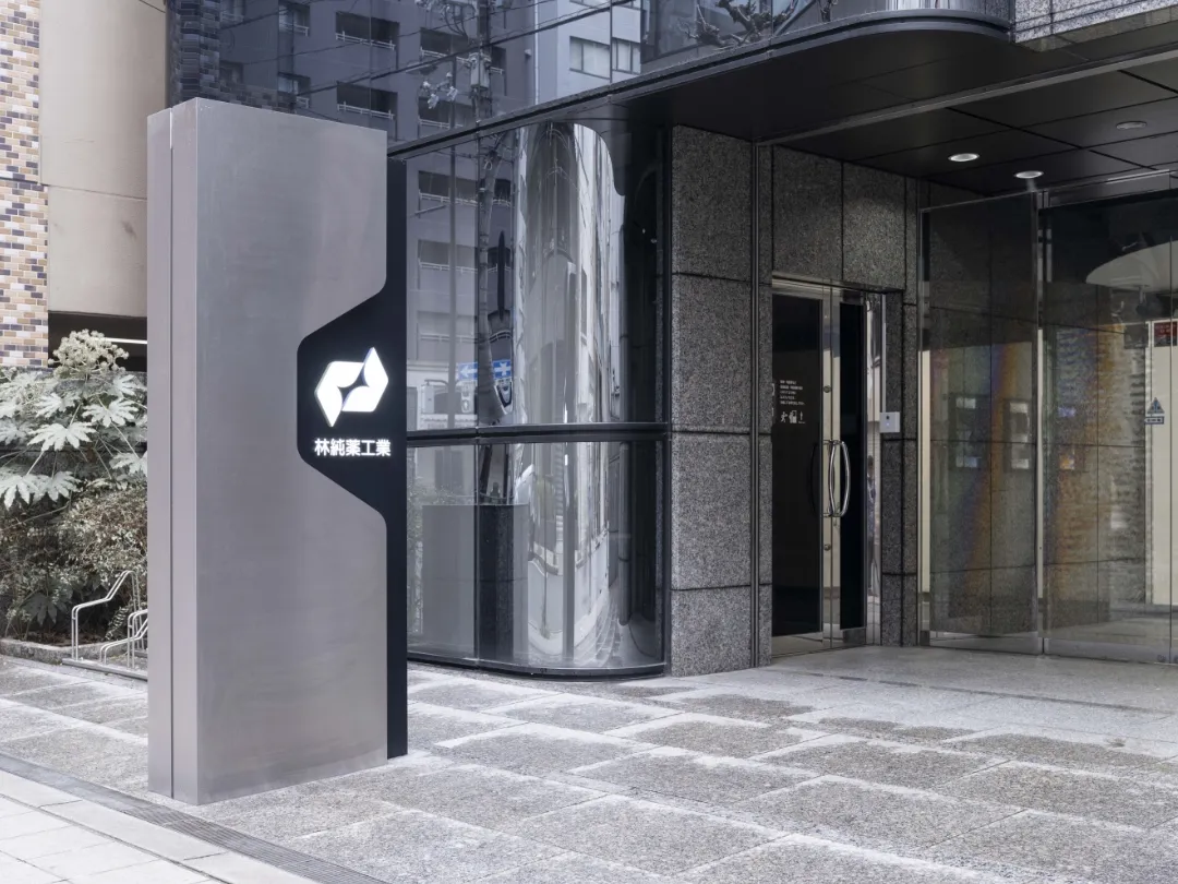 林純薬工業のロゴが入ったスタイリッシュな自立看板があるビルのエントランス。ガラス張りの外壁と石畳の地面