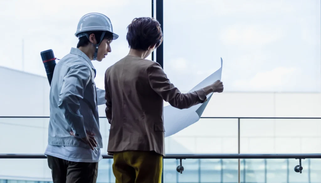 図面を見ながら窓際で打ち合わせをする作業着の男性とスーツの女性